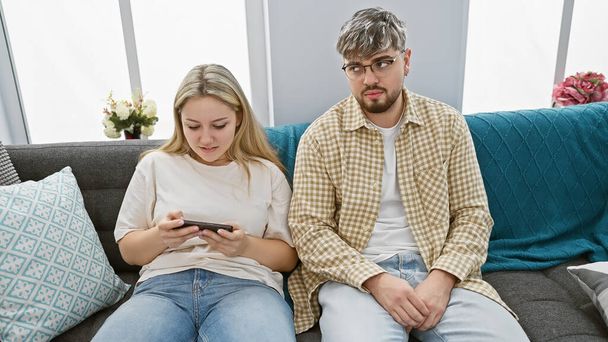 Jovem casal lounging no sofá com a mulher focada no smartphone e homem olhando para longe pensativo em um aconchegante apartamento interior. - Foto, Imagem