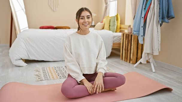 Una giovane donna ispanica sorride serena mentre seduta su un tappetino yoga nella sua camera da letto ordinata, irradiando benessere e comfort. - Foto, immagini