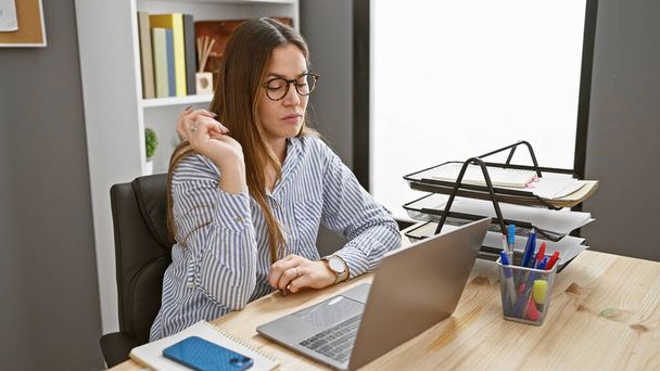 Una mujer reflexiva con gafas y camisa a rayas se sienta en su escritorio de la oficina trabajando en un ordenador portátil, evocando la esencia de un lugar de trabajo profesional moderno. - Foto, Imagen