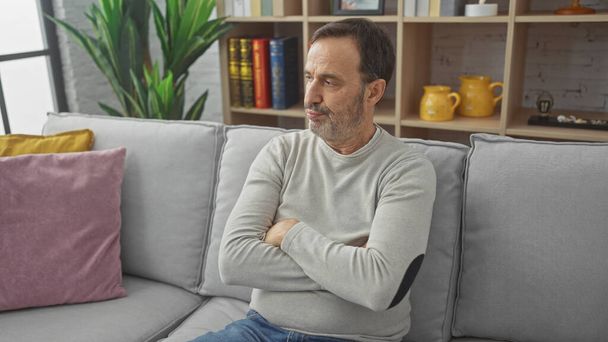 Egy elmélkedő középkorú, szakállas férfi ül keresztbe tett karokkal egy hangulatos nappaliban.. - Fotó, kép