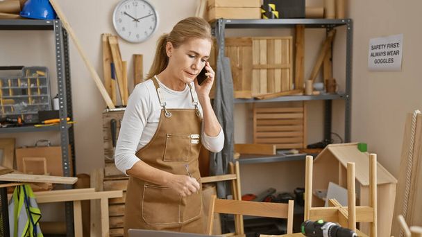 Μια γυναίκα με ποδιά μιλάει στο τηλέφωνο σε ένα καλά οργανωμένο εργαστήριο ξυλουργικής, που περιβάλλεται από ξύλινα ράφια και εργαλεία. - Φωτογραφία, εικόνα
