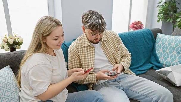 Ένα ζευγάρι χαλαρώνει σε έναν καναπέ με smartphones σε ένα άνετο σαλόνι, απεικονίζοντας τη σύγχρονη ζωή στο σπίτι. - Φωτογραφία, εικόνα