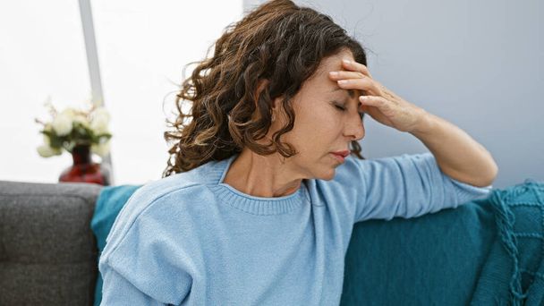 Una mujer hispana madura estresada con el pelo rizado toca su frente en el interior, expresando preocupación o dolor de cabeza en casa. - Foto, Imagen