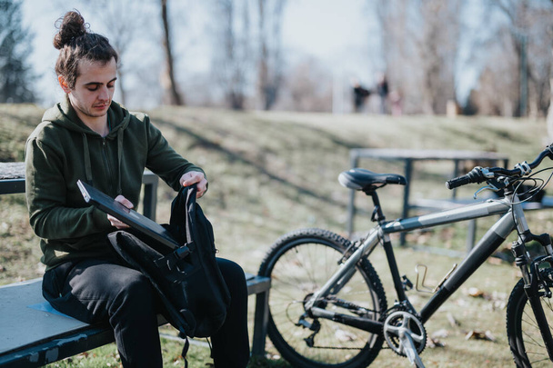 Nuori aikuinen mies rentoutuu puistossa, purkaa reppunsa istuessaan penkillä, ja hänen maastopyöränsä on pysäköity hänen viereensä aurinkoisena päivänä.. - Valokuva, kuva