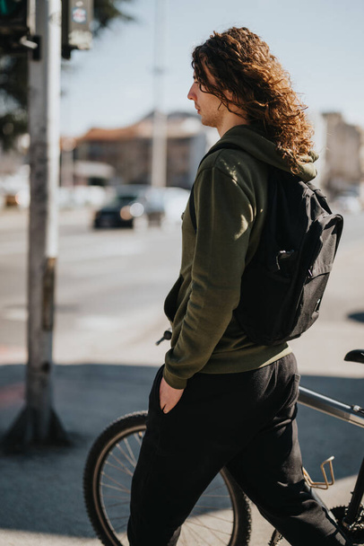 Розслаблений молодий чоловік з довгим кучерявим волоссям стоїть біля свого велосипеда, просочуючи сонце в ясний день, зображуючи відчуття свободи і міського дозвілля. - Фото, зображення