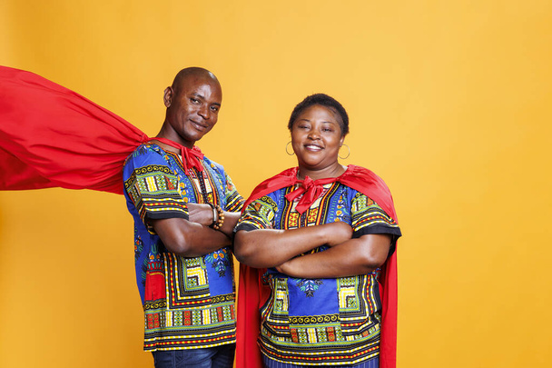 Smiling black couple dressed in superhero red cloaks posing with folded hands and looking at camera. Cheerful superman and superwoman pair wearing capes showing power and strength portrait - Photo, Image