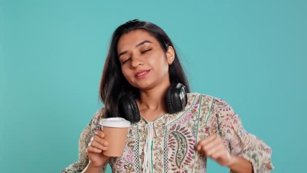 Mujer india alegre estiramiento y sorbiendo café fresco de la taza de papel desechable después de despertar. Persona animada radiante bebiendo bebida caliente temprano en la mañana, fondo del estudio, cámara B - Metraje, vídeo