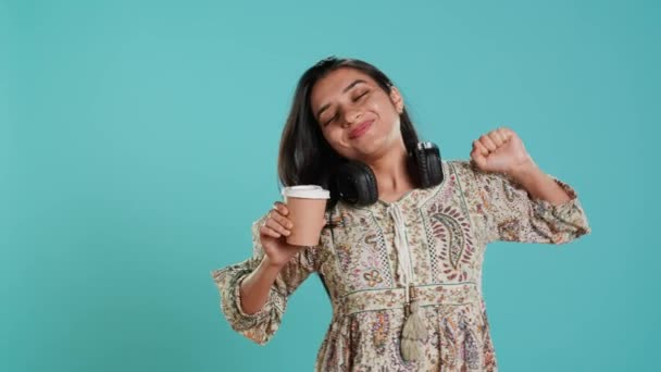 Mujer india alegre estiramiento y sorbiendo café fresco de la taza de papel desechable después de despertar. Persona animada radiante bebiendo bebida caliente temprano en la mañana, fondo del estudio, cámara A - Metraje, vídeo