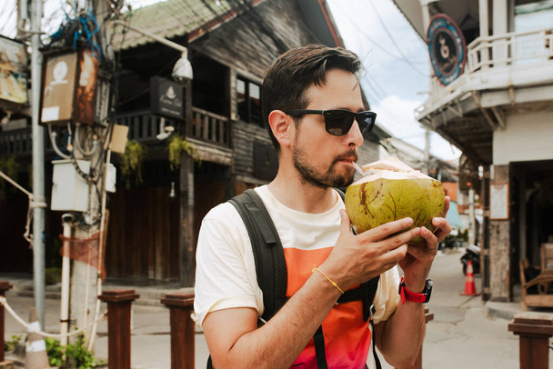Ένας άντρας με γυαλιά ηλίου απολαμβάνει φρέσκο νερό καρύδας στο γραφικό χωριό Fisherman, Koh Samui. Τα ρουστίκ ξύλινα κτίρια προσθέτουν στη γοητεία της τοποθεσίας. - Φωτογραφία, εικόνα