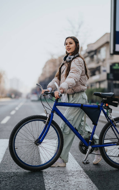 Modna młoda kobieta ze słuchawkami na szyi jeździ na rowerze po ulicy miejskiej, prezentując miejski styl życia i ekologiczny transport. - Zdjęcie, obraz