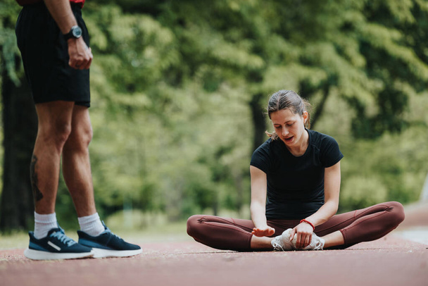 Motivierte junge Frau macht Strecken auf einer Laufstrecke, während ihr männlicher Trainer daneben steht, umgeben von üppigem Grün in einem ruhigen Park. - Foto, Bild