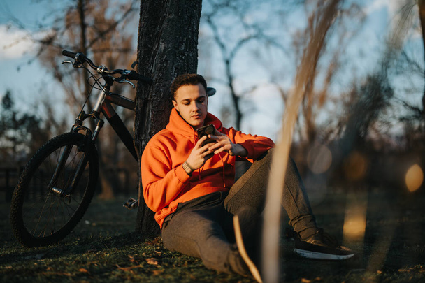 Ένας νεαρός άνδρας χαλαρώνει πάνω σε ένα δέντρο στο πάρκο, με ένα ποδήλατο βουνού δίπλα του, απολαμβάνοντας μια ήρεμη, ηλιόλουστη μέρα. - Φωτογραφία, εικόνα