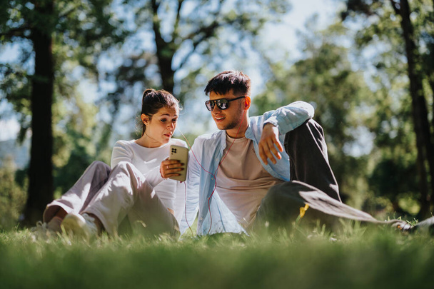 Um jovem casal se senta na grama em um parque ensolarado, compartilhando uma risada enquanto olha para um telefone inteligente. Esta imagem captura sua interação casual e alegre ao ar livre. - Foto, Imagem