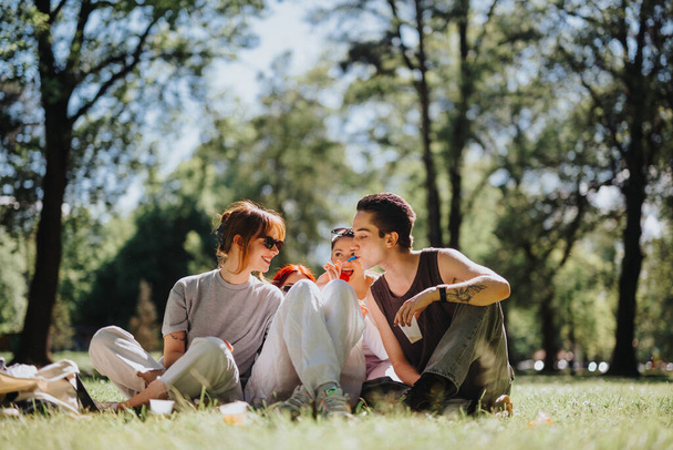 Kolme ystävää jakaa naurua ja ruokaa rauhallisen piknikin aikana rehevässä vihreässä puistossa nauttien auringon lämmöstä.. - Valokuva, kuva