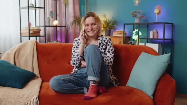 Smiling young Caucasian girl making phone conversation with friends sitting on sofa couch at home in room. Happy excited woman in casuals enjoying mobile talking gossiping at modern home apartment. - Footage, Video