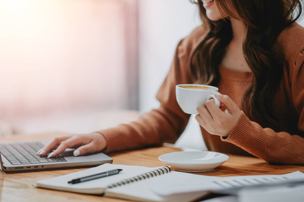 Close-up van de handen van een zakenvrouw die een koffiebeker vasthoudt terwijl ze op een laptop typt, wat een productief begin van de dag aangeeft. - Foto, afbeelding