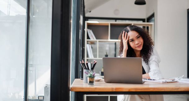 Προβληματική επιχειρηματίας με το χέρι στο μέτωπό της κάθεται στο laptop της σε έναν ηλιόλουστο, σύγχρονο χώρο γραφείου. - Φωτογραφία, εικόνα