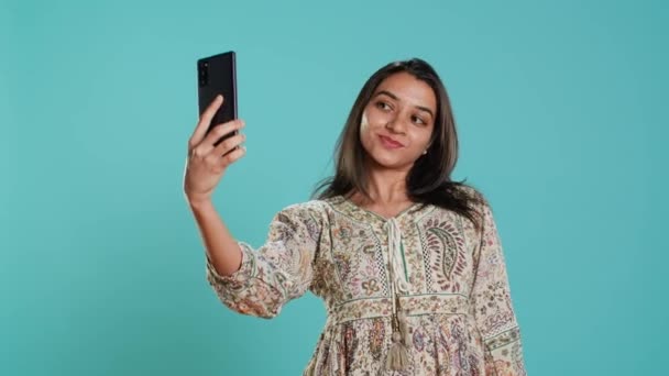 Vidám indiai nő okostelefont használ, hogy szelfiket készítsen és posztolja őket a közösségi médián. Happy személy fotózás segítségével telefon szelfi kamera, elszigetelt stúdió háttér, kamera A - Felvétel, videó