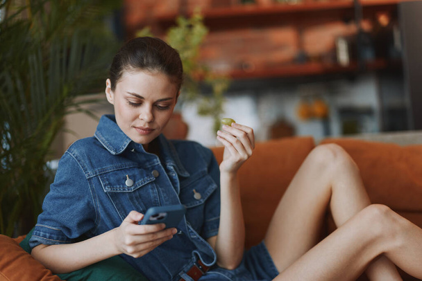 Nuori nainen istuu sohvalla ristissä jalat, katsellen matkapuhelin näytön tarkkaavaisesti - Valokuva, kuva