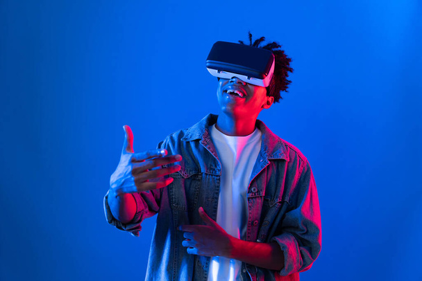 Νεαρός Αφροαμερικανός που φοράει VR δείχνοντας το άδειο διαστημικό προϊόν που λανσάρει το εμπορικό σήμα εξηγώντας λεπτομέρειες για μπλε ροζ νέον φως τοίχο φουτουριστική τεχνολογία εικονική πραγματικότητα σε metaverse κόσμο. Συμβολή. - Φωτογραφία, εικόνα