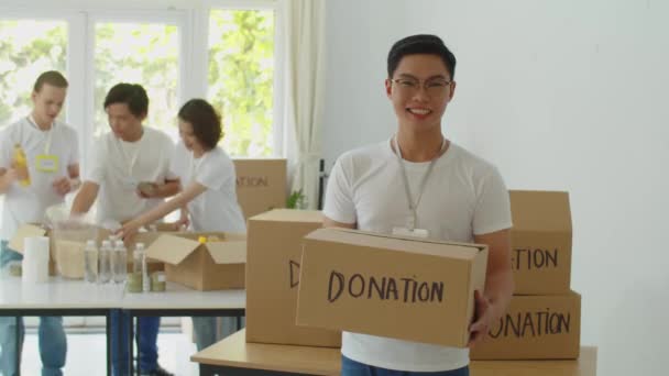 Retrato mediano del alegre hombre asiático voluntario en el centro de donación y sonriendo a la cámara - Imágenes, Vídeo