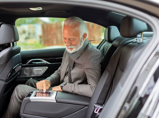 Distinguido hombre mayor en un traje ajusta el panel de control de pantalla táctil de un vehículo de lujo mientras viaja. - Foto, Imagen