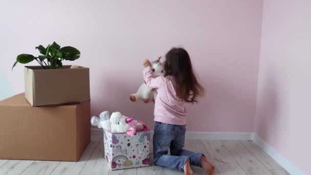 Маленькая девочка достает игрушки из коробки в его новой комнате в новом доме. Маленькая девочка счастлива переехать в свою новую комнату. Новое жилье. Недвижимость и аренда, кредит и ипотека, день переселения - Кадры, видео