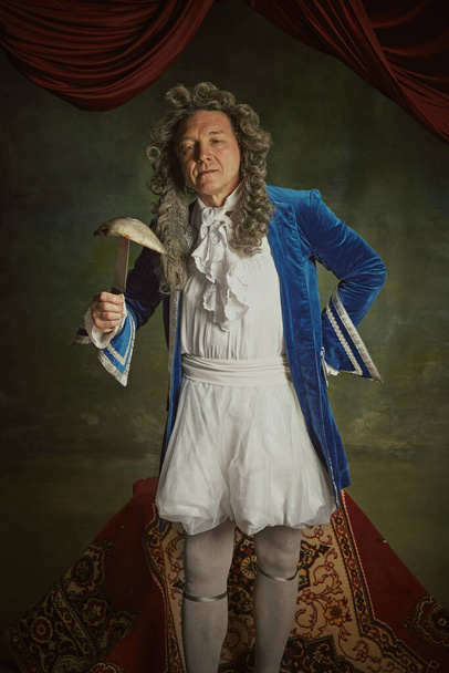 Portrait of elderly man dressed in richly detailed baroque costume posing holding fish on fork against vintage studio background. Concept of comparisons of eras, fusion of modernity and history. - Photo, Image