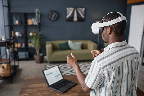 Молодий афроамериканський чоловік у гарнітурі VR, що стоїть у вітальні, серфінг Інтернет на ноутбуці, копіювати простір - Фото, зображення