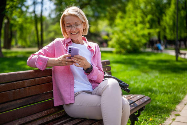 Πορτρέτο της ευτυχισμένης ώριμης γυναίκας στο πάρκο. Στέλνει μηνύματα στο τηλέφωνο.. - Φωτογραφία, εικόνα