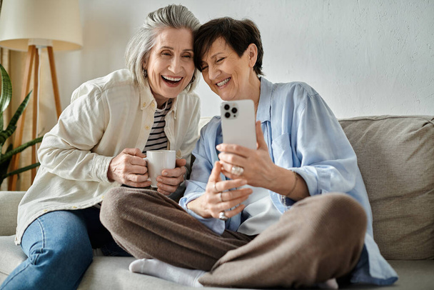 Zwei ältere Frauen sitzen auf einer Couch und machen fröhlich zusammen ein Selfie. - Foto, Bild