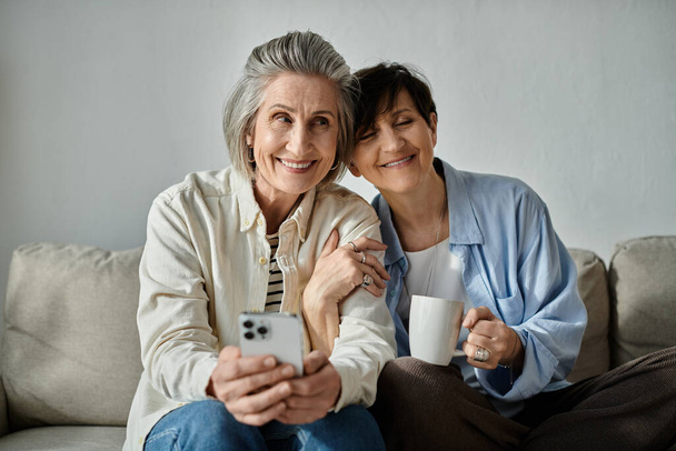 Dwie kobiety, kochająca dojrzała lesbijska para, siadają na kanapie, pochłonięte telefonem komórkowym. - Zdjęcie, obraz