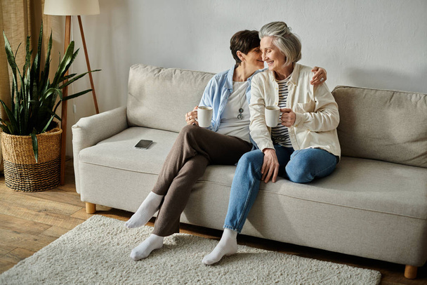 Dwie starsze osoby, kochająca dojrzała para lesbijek, siadają razem na kanapie, delektując się filiżankami kawy. - Zdjęcie, obraz