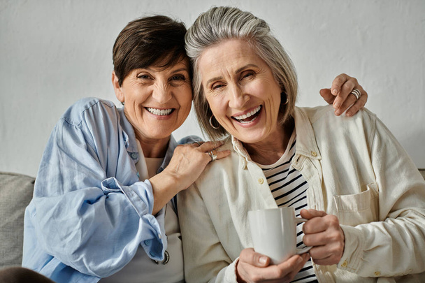 Dwie starsze kobiety, kochająca dojrzała para lesbijek, siadają razem na kanapie, delektując się filiżanką kawy. - Zdjęcie, obraz