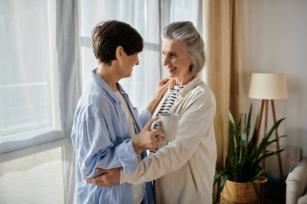 Δύο ηλικιωμένες γυναίκες μοιράζονται μια εγκάρδια αγκαλιά από ένα ηλιόλουστο παράθυρο.. - Φωτογραφία, εικόνα
