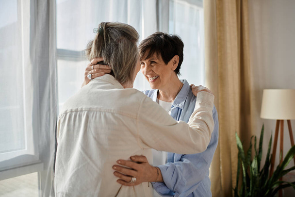 Δύο ηλικιωμένες γυναίκες μοιράζονται μια εγκάρδια αγκαλιά μπροστά από ένα φωτεινό παράθυρο. - Φωτογραφία, εικόνα