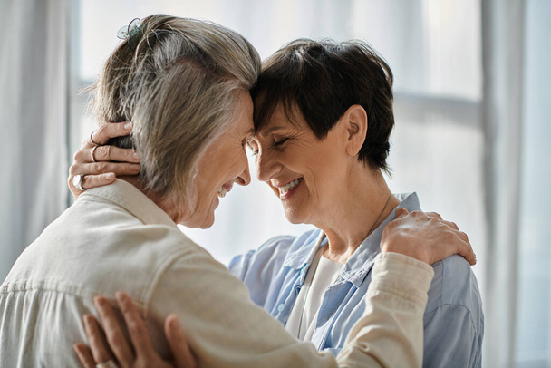 Two senior women hug warmly in front of a bright window. - Photo, Image