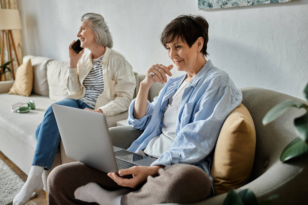 Két nő, egy szerető érett leszbikus pár, telefonon beszélgetnek és laptopot használnak a kanapén.. - Fotó, kép