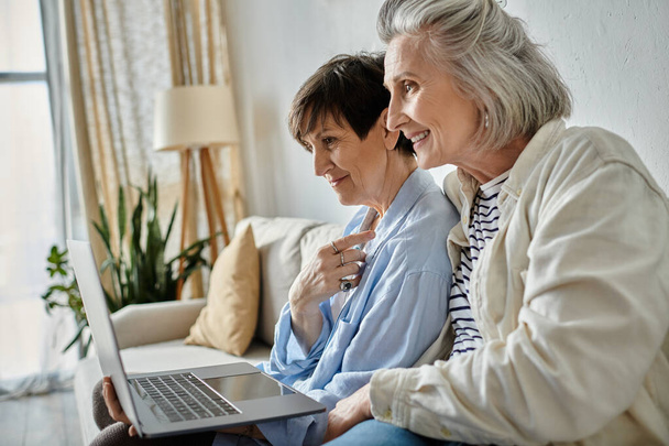 Δύο μεγαλύτερες γυναίκες απολαμβάνουν τη χρήση ενός φορητού υπολογιστή, ενώ κάθεται σε έναν καναπέ μαζί. - Φωτογραφία, εικόνα