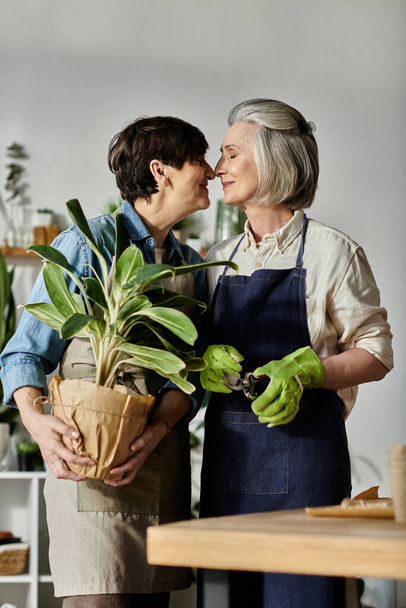 Two women in aprons kiss in front of potted plant. - Photo, Image