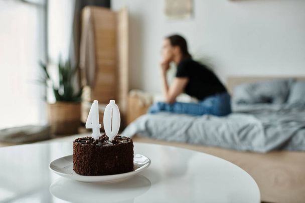 A middle-aged woman sits alone on a bed with a cake, lost in thought. - Photo, image