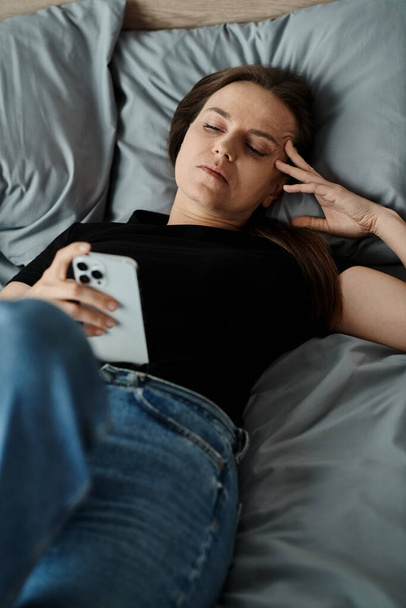 Woman lays in bed with phone on head. - Фото, зображення