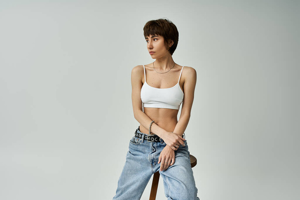 Young woman in jeans and white top sits calmly on a stool. - Φωτογραφία, εικόνα