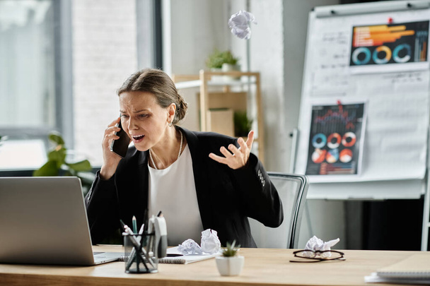 Middle-aged woman talking on phone at office desk, overwhelmed by stress. - Foto, Bild