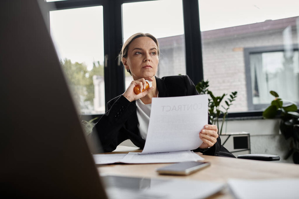 Middle aged woman sitting at desk, reading paper with a concerned expression. - Photo, image