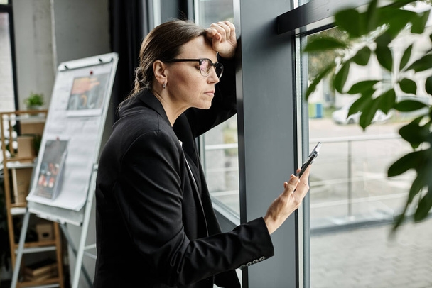 Middle-aged woman in glasses leans against window, looking at phone screen. - Photo, Image
