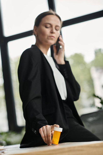 Middle-aged woman in office, multitasking with phone and pill bottle. - Fotografie, Obrázek