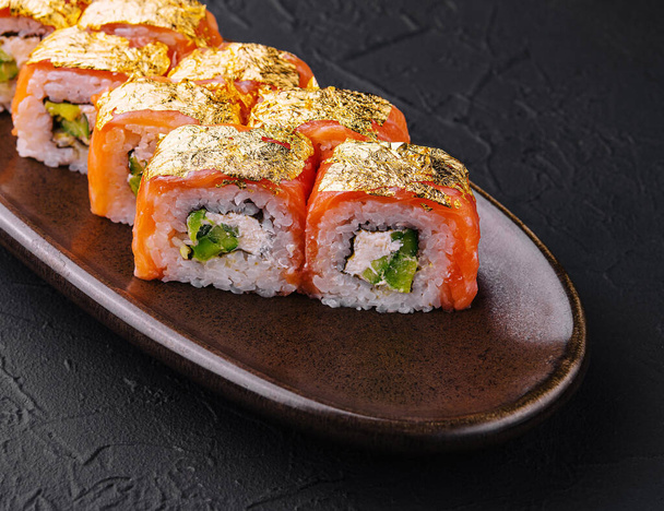 Elegáns sushi tekercs tetején arany levél, szolgált kerámia edényben ellen sötét texturált háttér - Fotó, kép