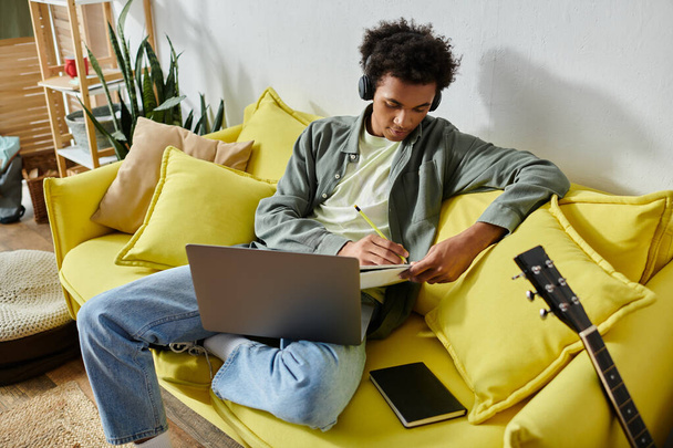 A young African American man sits on a yellow couch with headphones and a laptop. - Fotoğraf, Görsel