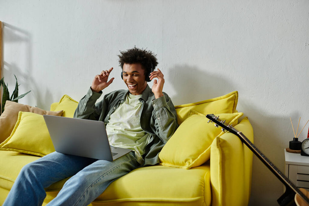 Young man engrossed in online studying with headphones and laptop on yellow couch. - Φωτογραφία, εικόνα